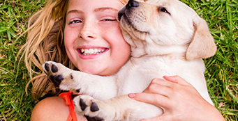 A blonde girl holds a yellow lab puppy while lying on her back in the grass.